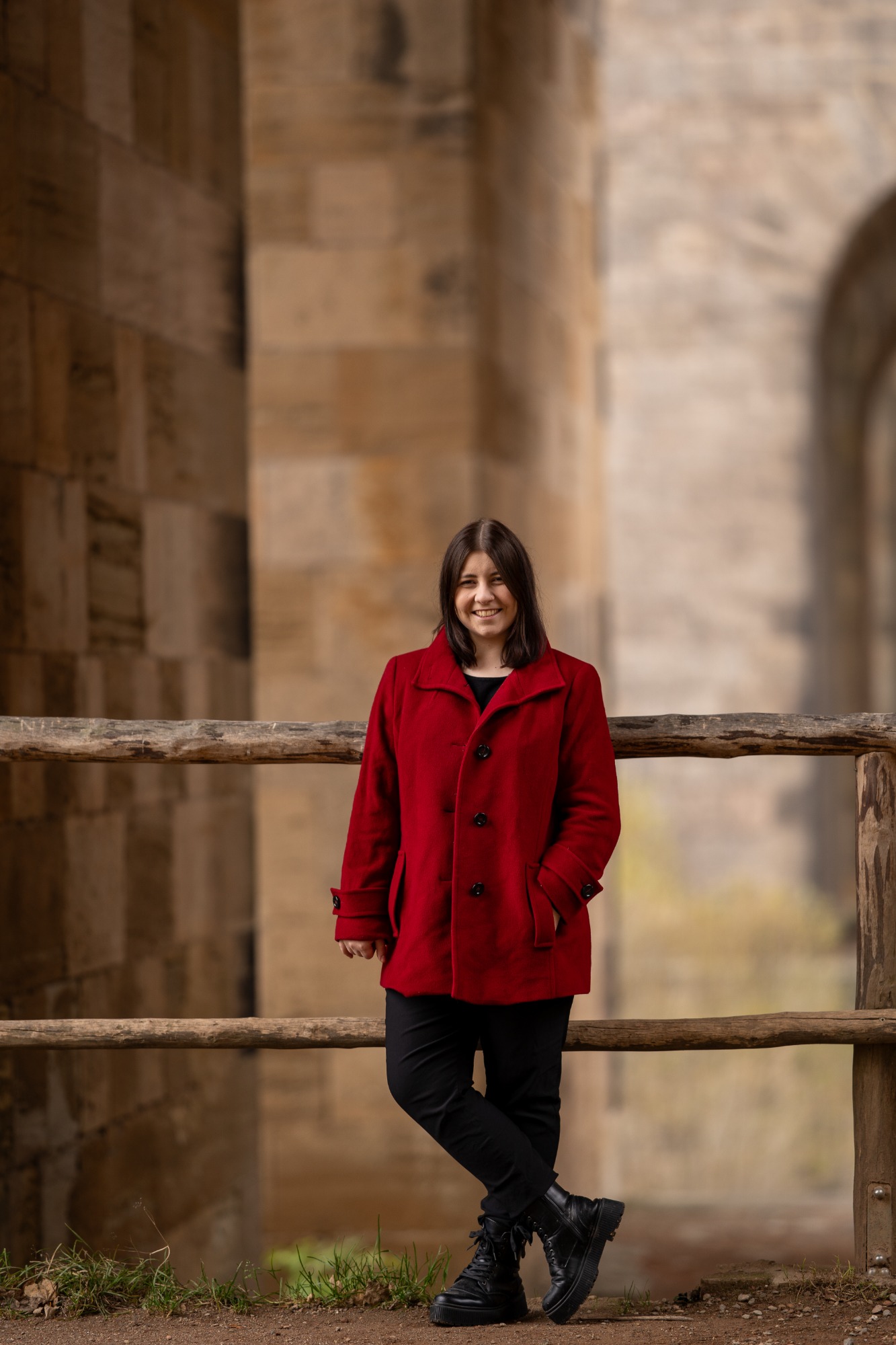 Portrait Frau in rotem Mantel steht lächelnd an einem Holzgeländer vor einer Steinmauer.